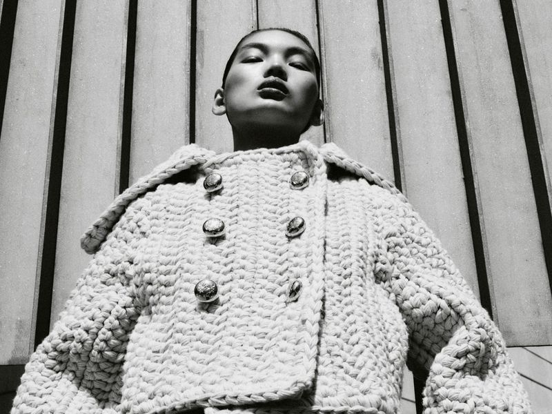 Low-angle shot of a woman in a textured jacket with metallic buttons, against a striped background, black and white, fashion editorial.