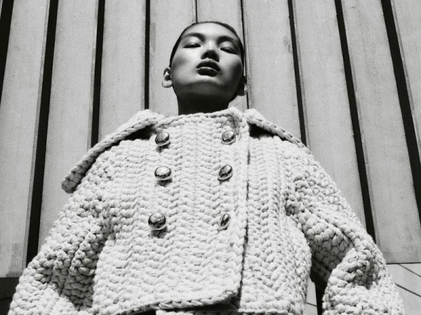Low-angle shot of a woman in a textured jacket with metallic buttons, against a striped background, black and white, fashion editorial.