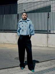 Model wearing a light blue textured cropped jacket and dark trousers standing outdoors against a metal fence