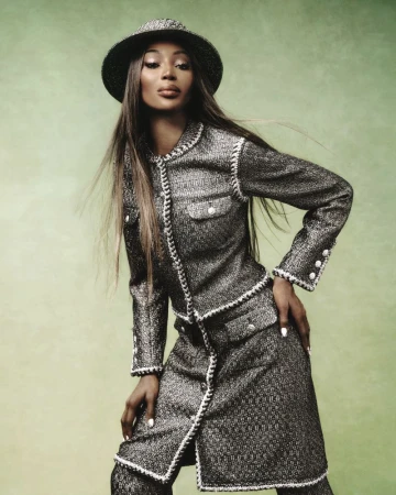 Elegant adult female fashion model poses in a tweed skirt suit with a matching hat against a green backdrop in a studio portrait.