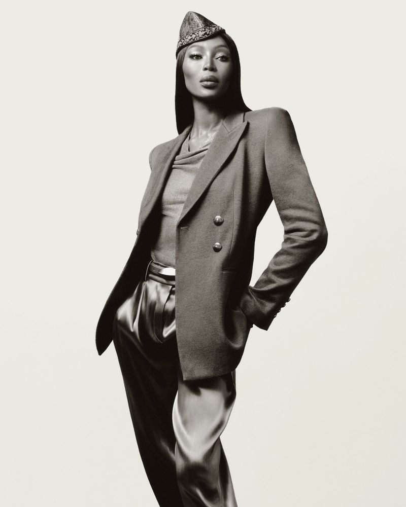 Black and white fashion portrait of an adult woman wearing a blazer, satin pants, and a decorative hat in a photo studio setting.