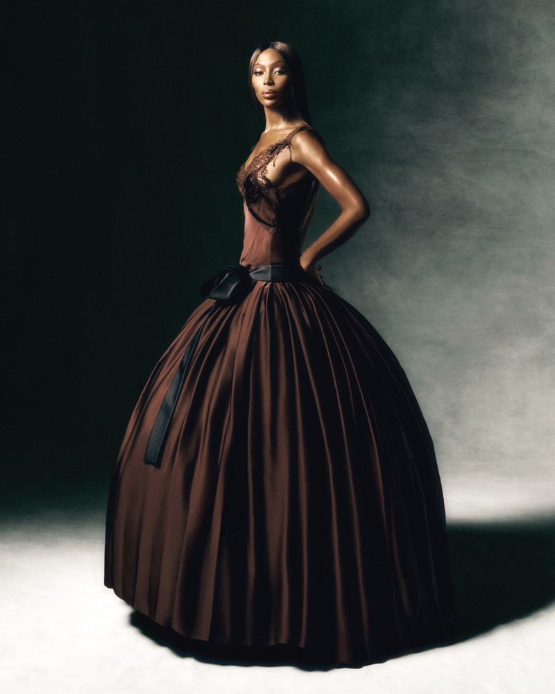 Woman posing in a brown ballgown with a black bow, wearing lace details, in a studio with dramatic lighting and dark background.