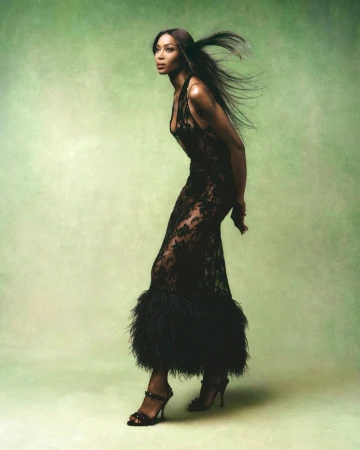 A full-length portrait of a Black female fashion model in a lace gown with black feather trim, posing against a light green background.