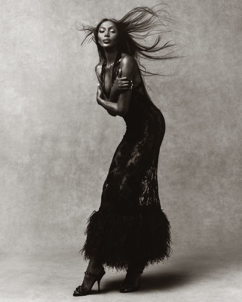 Black and white portrait of a woman in a flowing black gown, hair blowing, set against a neutral backdrop in a photo studio.
