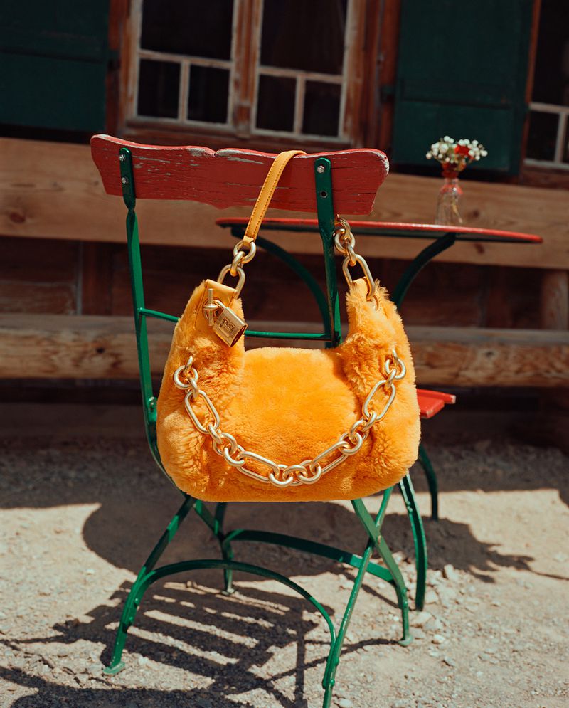 Orange furry purse with a gold chain on a green metal chair in front of a wood building with green shutters.