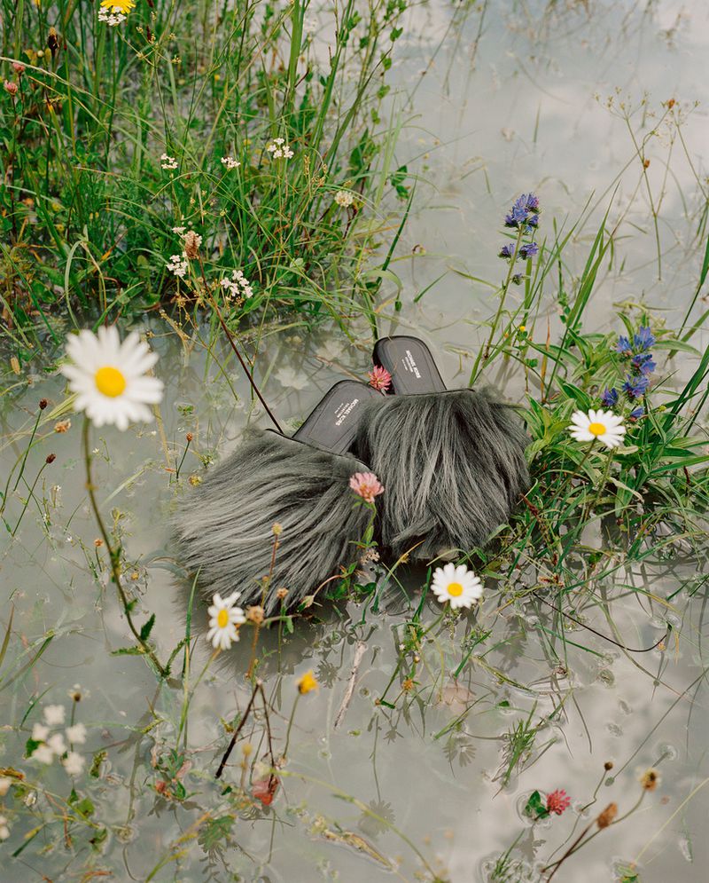 Grey, furry slide sandals in a shallow pool of water surrounded by daisies and wildflowers in a bright, outdoor setting.