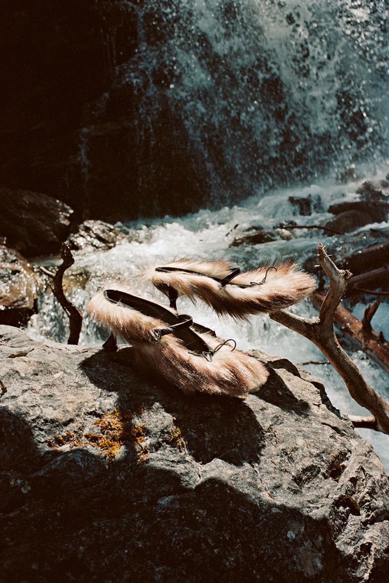 Two furry loafers sit on a rock with a waterfall in the background, showcased in an outdoor environment with natural lighting.