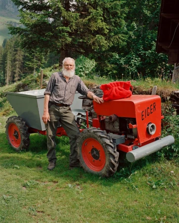 An elderly man with a white beard stands by a small red tractor with gray cargo trailer on a grassy hill in a rural setting.