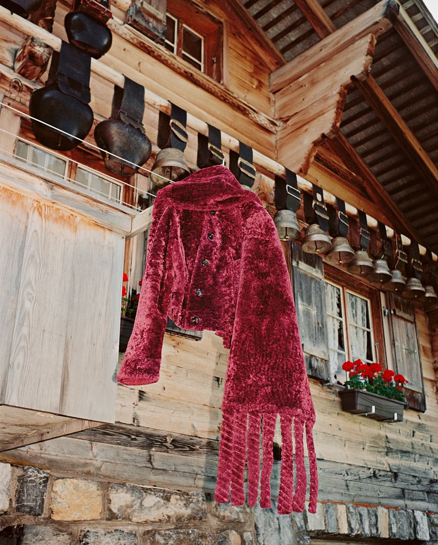 Maroon fringed coat on rustic wood building exterior with window boxes filled with bright red geraniums, decorative metal bells.