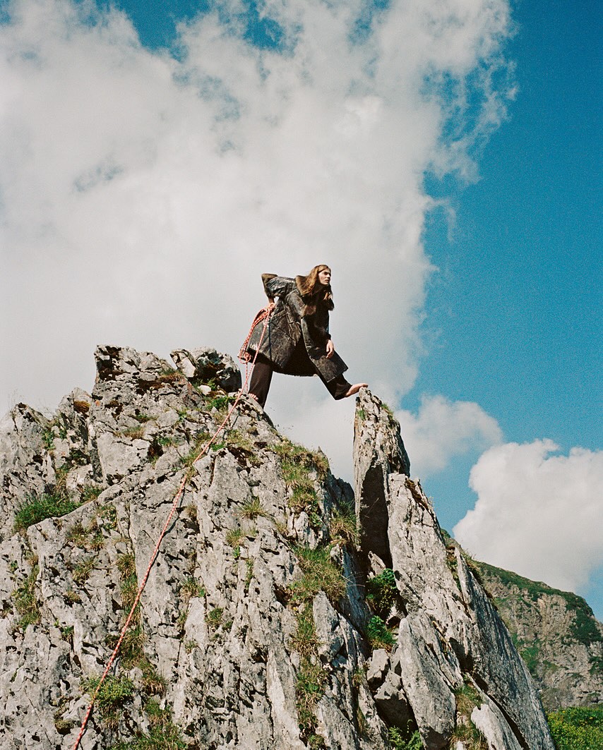 Adult female posing on a rocky mountain peak wearing a coat, with blue sky and clouds, photographed at eye-level in an outdoor setting.