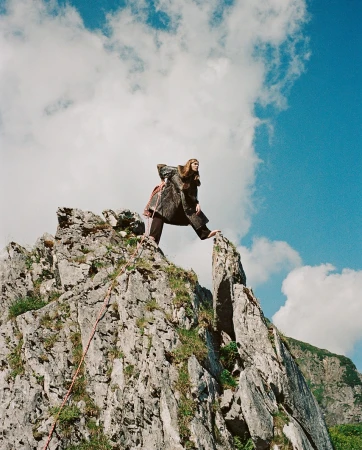 Adult female posing on a rocky mountain peak wearing a coat, with blue sky and clouds, photographed at eye-level in an outdoor setting.