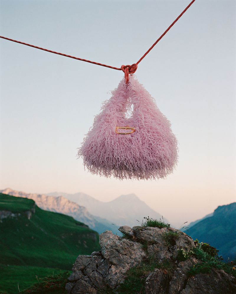 Fluffy lavender handbag suspended by a red rope against a backdrop of green mountains and a blue sky at daytime.