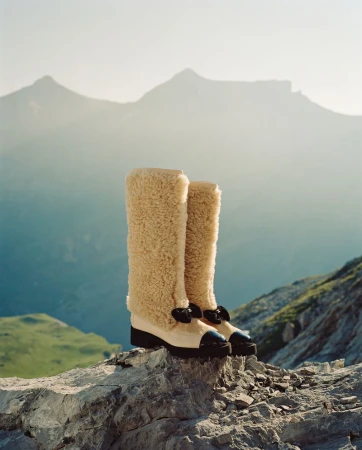 Fashionable shearling boots displayed on a rocky surface with a mountain range backdrop under soft, natural light.