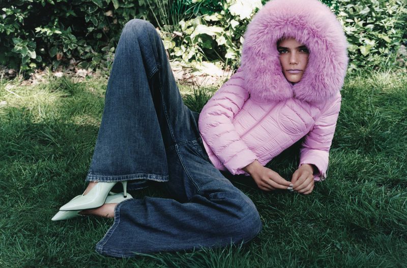 A woman in jeans and pale green heels lies in the grass, wearing a pink puffer jacket with a fur hood, in an outdoor fashion shot.