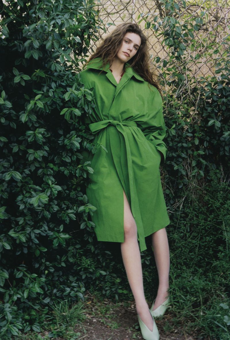 An adult woman with long hair in a green trench coat poses outdoors next to a leafy bush and chain link fence, looking at the camera.