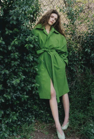 An adult woman with long hair in a green trench coat poses outdoors next to a leafy bush and chain link fence, looking at the camera.