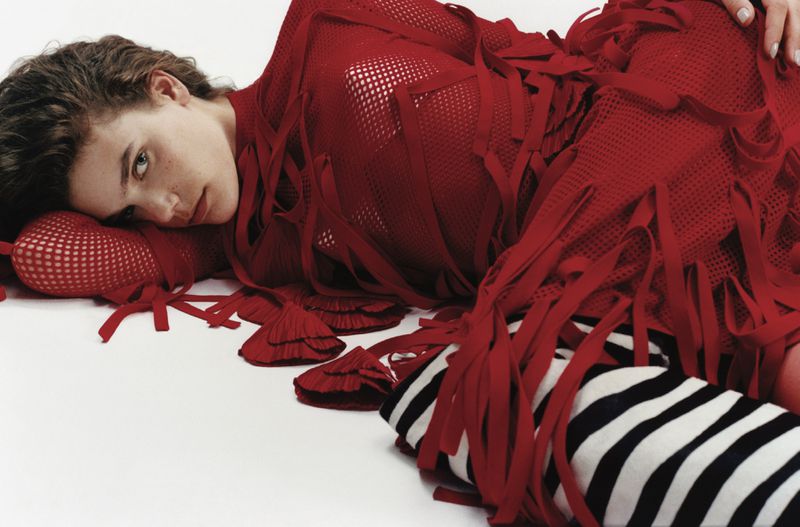 A young woman with wavy brunette hair and blue eyes lies on a white surface wearing a red mesh dress and black and white leggings.