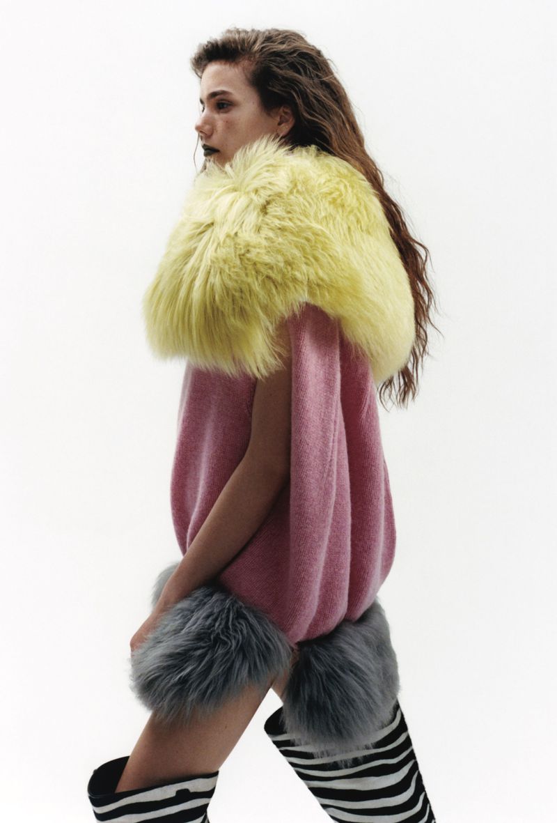 A person poses in a studio wearing a pink sweater with yellow fur collar, striped boots, and gray fur bands on arms. Profile shot.
