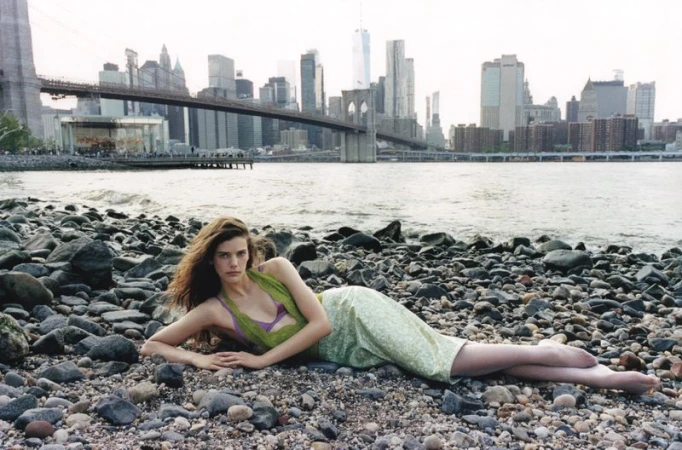 Woman in a green dress poses on a rocky beach with the Brooklyn Bridge and New York City skyline in the background.