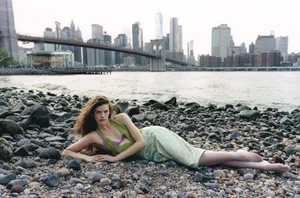 Model lying on a rocky shore with the Manhattan skyline and a suspension bridge across the water in the background.