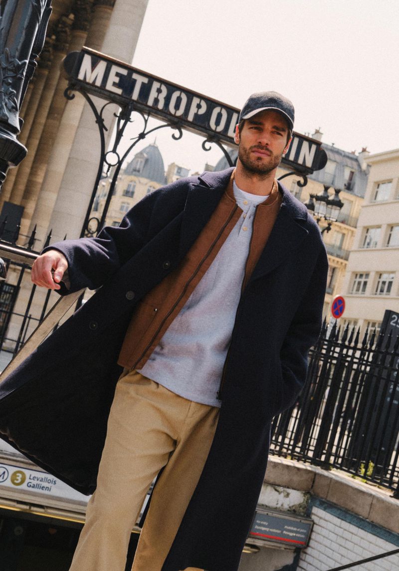 An adult male model poses in a dark coat and cap in front of a historic Métropolitain subway entrance in an urban setting.