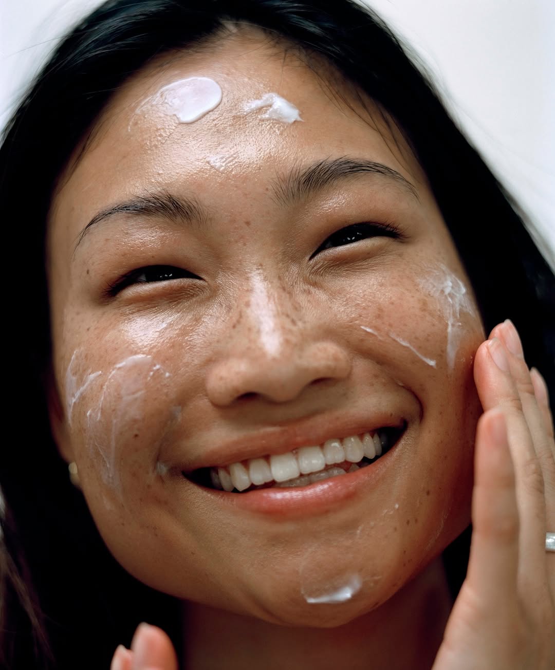 Close-up of a smiling Asian woman applying white cream to her face and forehead in an indoor beauty or skincare setting.