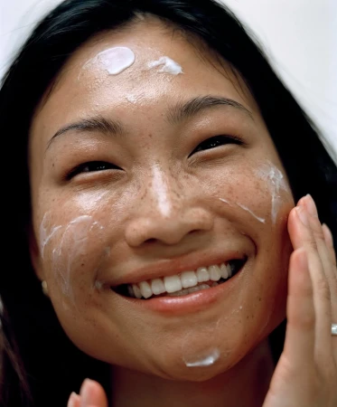Close-up of a smiling Asian woman applying white cream to her face and forehead in an indoor beauty or skincare setting.