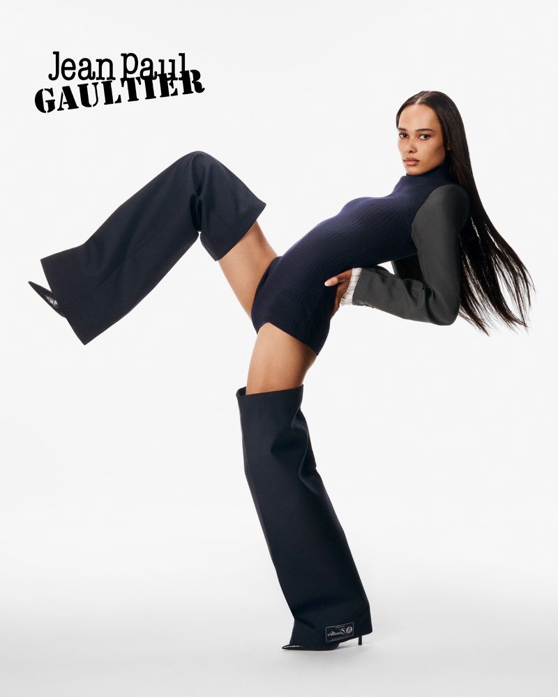 Fashion editorial shot of a posing adult female wearing a dark bodysuit and unusual pants, against a bright white backdrop in studio lighting.