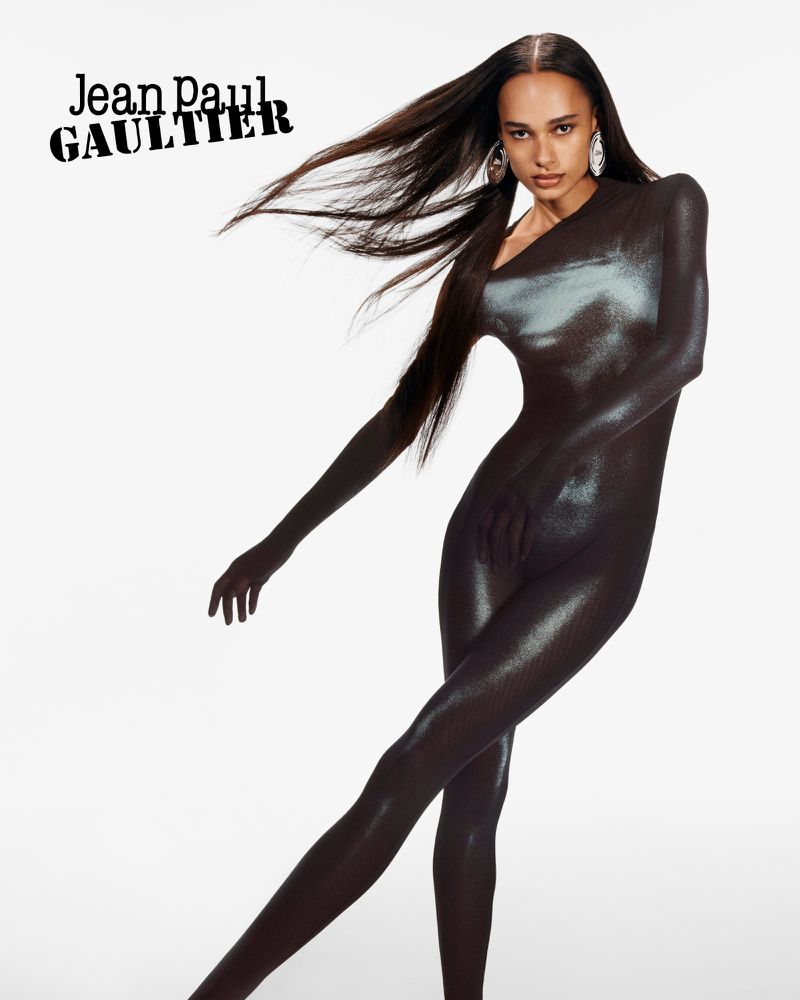 A woman in a metallic black catsuit strikes a pose on a white backdrop, with long, flowing hair and large, reflective earrings.