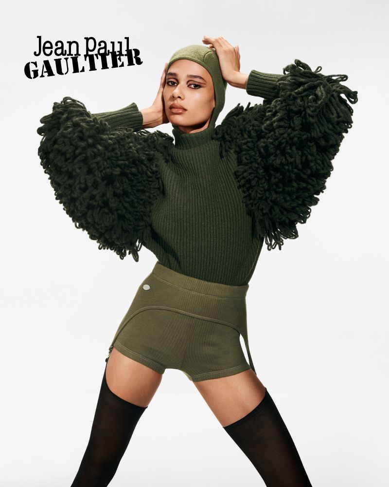 A woman with dark hair poses in a green sweater, shorts, hat, and black tights against a white backdrop with Jean Paul Gaultier text.