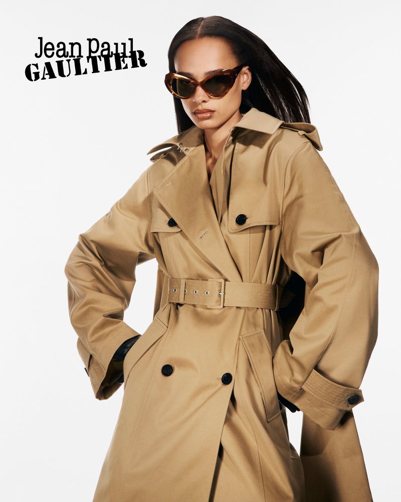 An adult female model poses in a tan belted trench coat and sunglasses in front of a white background. Designer Jean Paul Gaultier logo visible.