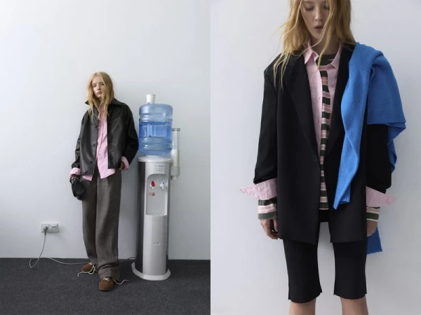 Fashionable woman in black jacket and brown trousers stands by a water cooler in a minimalist setting, featuring a stylish outfit.