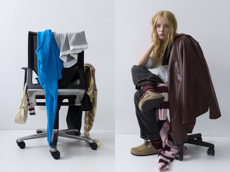 Indoor shot of an adult female sitting on a chair next to a chair with clothing draped over it in front of a white wall.