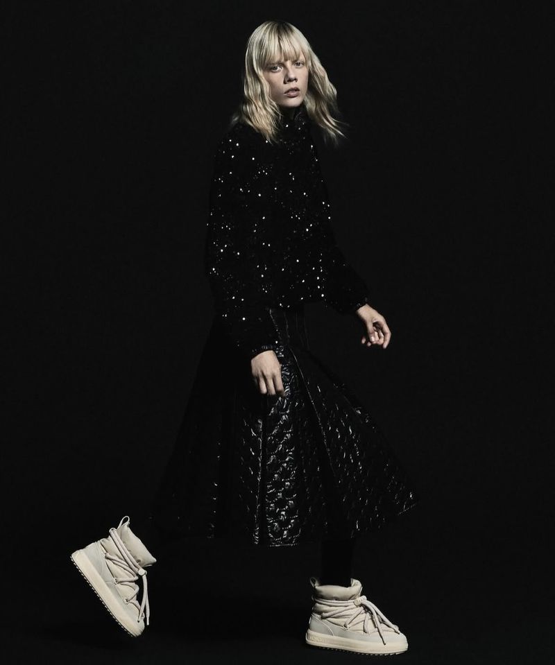 Fashionable adult female strides confidently in a black sequined top, black quilted skirt, and beige sneakers against a dark backdrop.