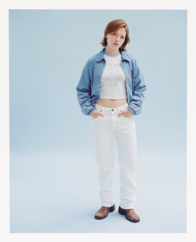 Woman in denim jacket and white jeans poses in a studio with blue background, showcasing casual fashion and youthful style.