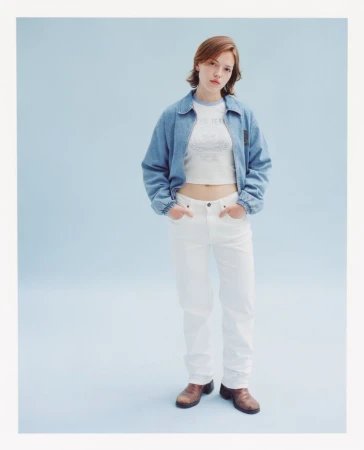 Woman in denim jacket and white jeans poses in a studio with blue background, showcasing casual fashion and youthful style.