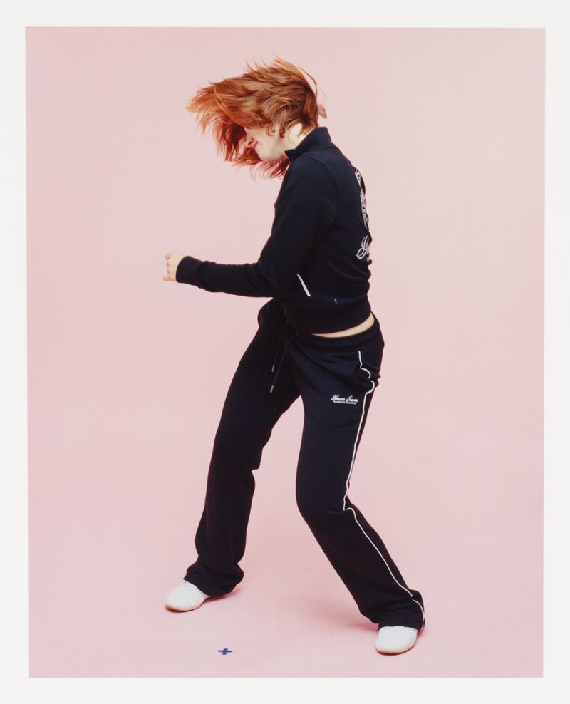 A young woman with red hair dances in a black tracksuit against a light pink backdrop, illuminated by soft lighting in a photo studio.