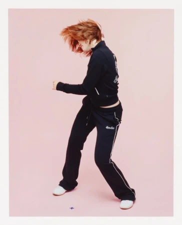 A young woman with red hair dances in a black tracksuit against a light pink backdrop, illuminated by soft lighting in a photo studio.