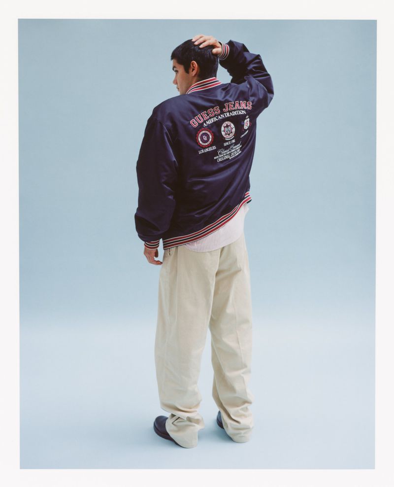 Rear shot of a male wearing a blue GUESS jeans bomber jacket with tan pants standing in a light blue photo studio, fashion shot.