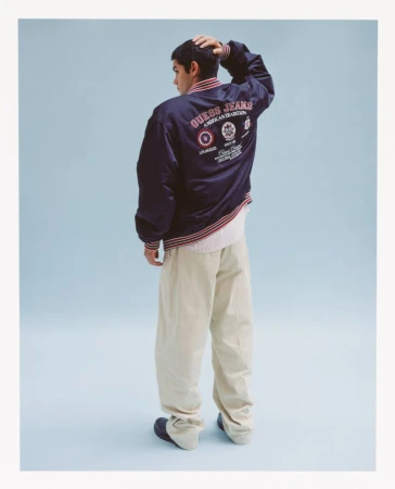 Rear shot of a male wearing a blue GUESS jeans bomber jacket with tan pants standing in a light blue photo studio, fashion shot.