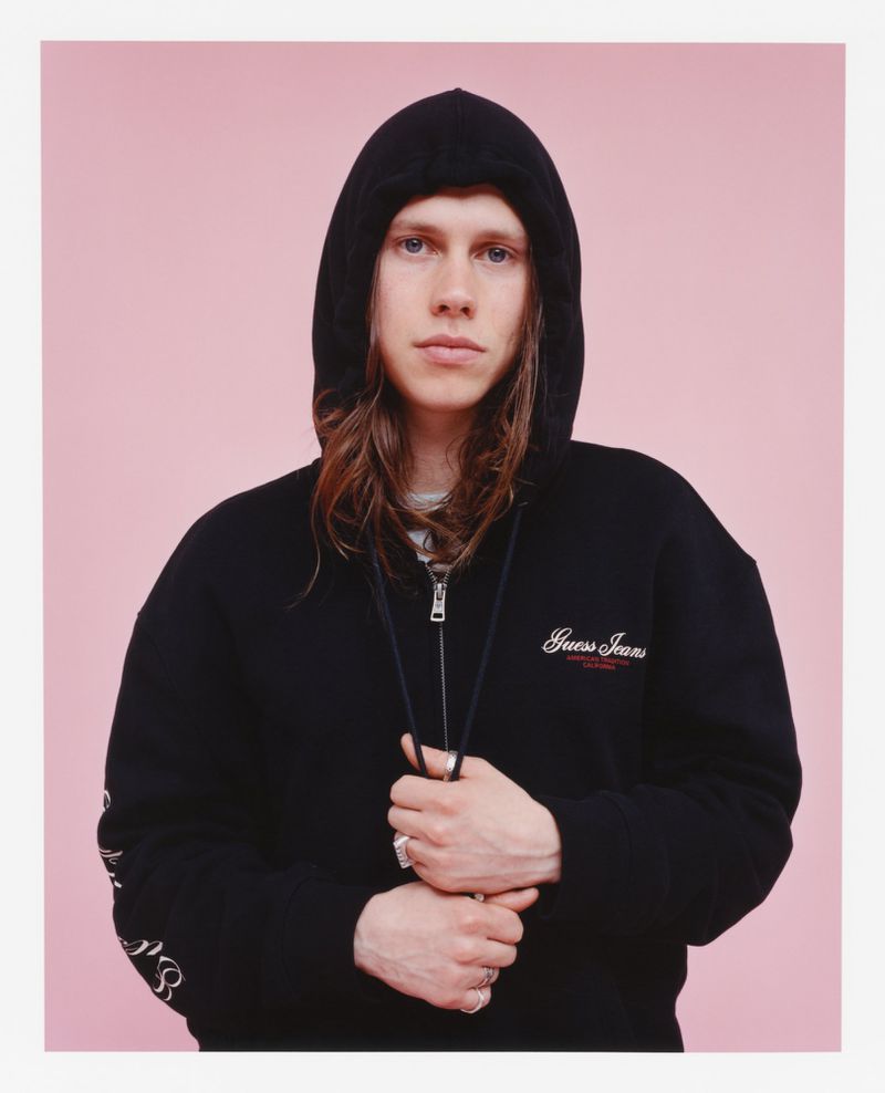 Portrait of a young adult with long brown hair wearing a black hooded sweatshirt against a pink background in a studio shot.
