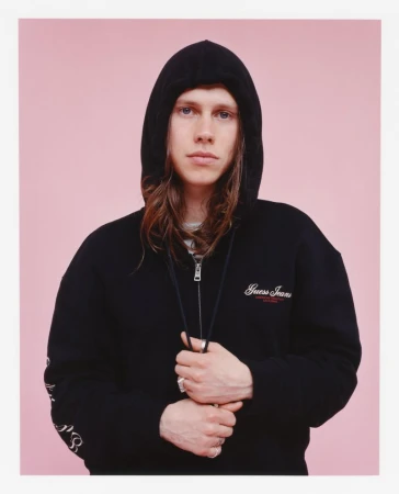 Portrait of a young adult with long brown hair wearing a black hooded sweatshirt against a pink background in a studio shot.