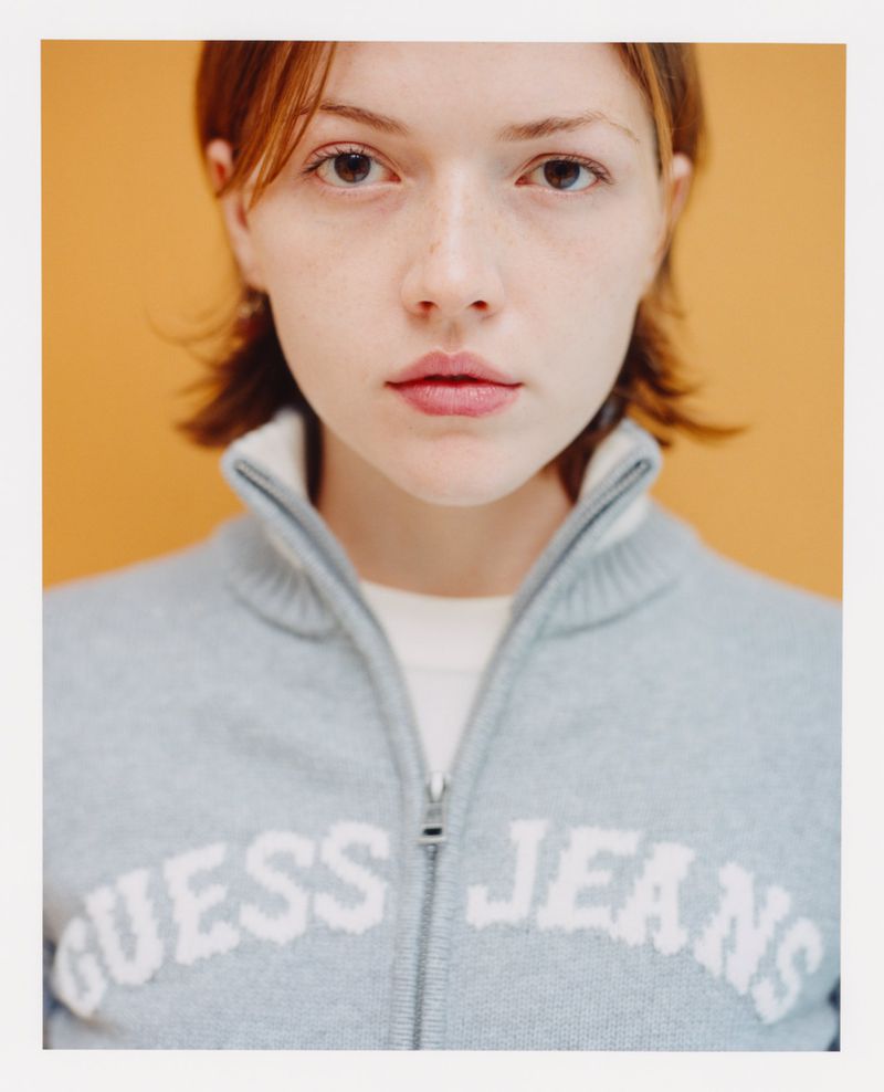 Close-up portrait of a woman with auburn hair wearing a gray zip-up sweatshirt with white 'GUESS JEANS' logo, set against an orange background.
