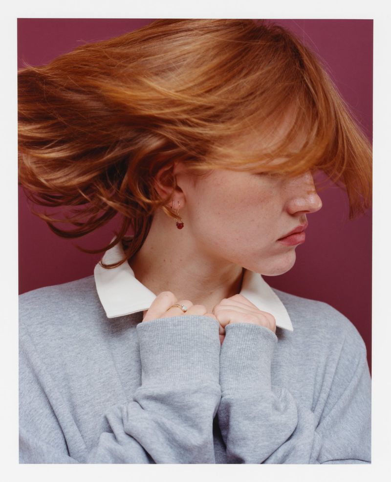 Close-up portrait of a young woman with red hair and grey sweater, posing indoors against a purple background in a natural lighting style.