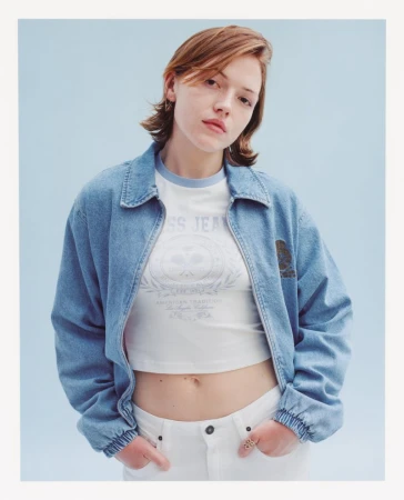 Woman with short reddish-brown hair wearing a light blue denim zip jacket, white cropped t-shirt, and white pants against a pale blue background.