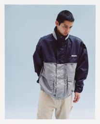 A man wearing a navy and grey colorblock zip jacket with beige trousers standing against a pale blue studio background.