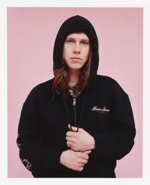 Portrait of a man with long hair wearing a black hooded zip sweatshirt against a solid light pink backdrop.