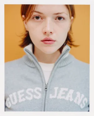 Close-up portrait of a woman wearing a light blue Guess Jeans zip-up sweater against a solid warm yellow background.