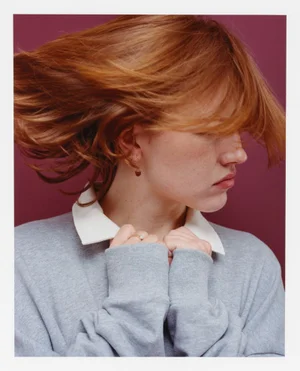 Close-up portrait of a red-haired woman wearing a grey sweatshirt with white collar against a dark red backdrop.