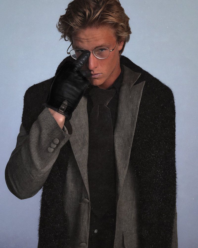 Portrait of a young adult man wearing glasses, gloves, a black tie, and a gray and black coat in front of a light blue background.